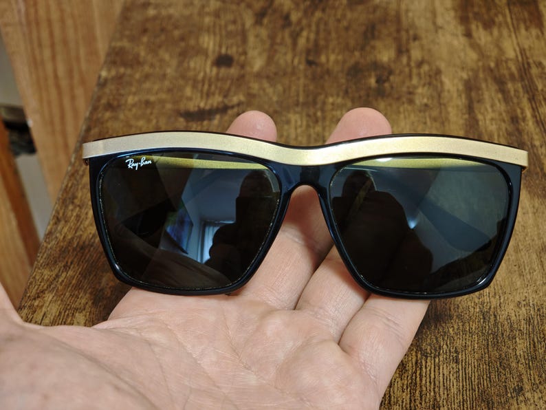 Peut inclure: Lunettes de soleil noires avec une monture sup&eacute;rieure dor&eacute;e. Les lunettes ont des verres fonc&eacute;s et le logo Ray-Ban est visible sur la lentille gauche. Les lunettes sont tenues dans une main.