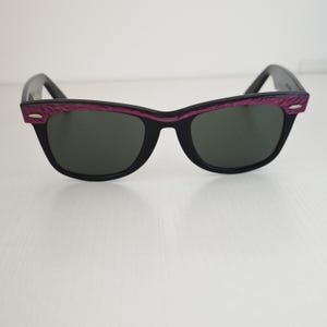 Puede incluir: Gafas de sol con montura negra y lentes de color verde oscuro. La parte superior del marco presenta un acabado texturizado en color morado intenso con peque&ntilde;os detalles plateados en las esquinas exteriores. Dise&ntilde;o cl&aacute;sico con patillas.