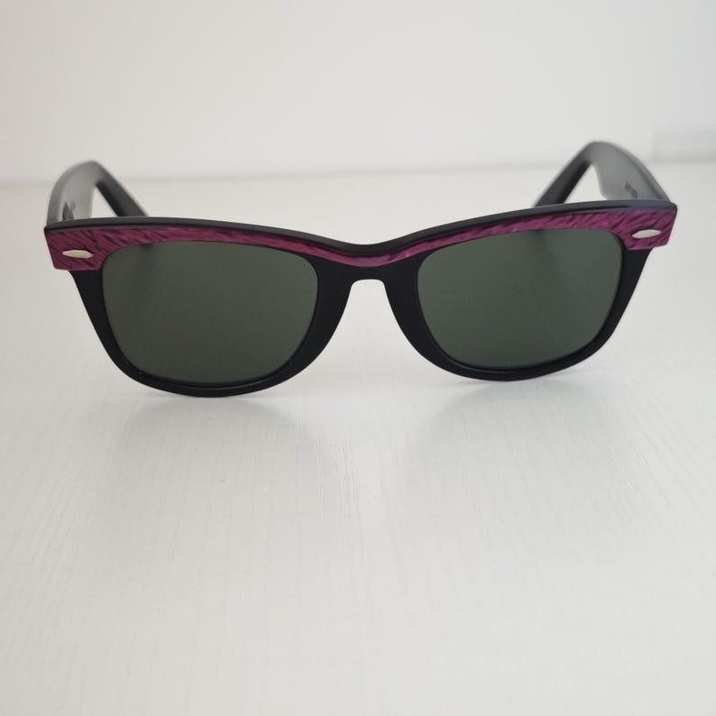 Puede incluir: Gafas de sol con montura negra y lentes de color verde oscuro. La parte superior del marco presenta un acabado texturizado en color morado intenso con peque&ntilde;os detalles plateados en las esquinas exteriores. Dise&ntilde;o cl&aacute;sico con patillas.