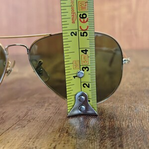 May include: Gold-framed aviator sunglasses with brown lenses. The sunglasses are measured with a yellow tape measure, showing a height of approximately 2 inches. The glasses are on a wooden surface.