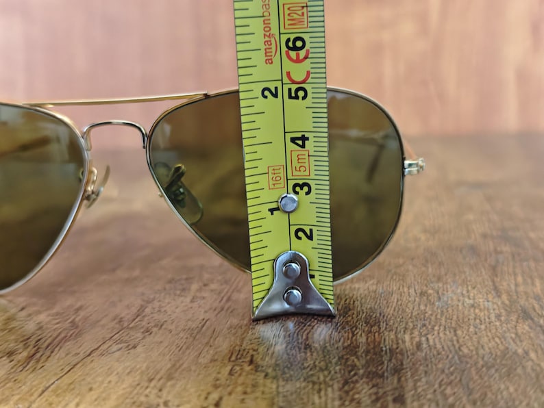 May include: Gold-framed aviator sunglasses with brown lenses. The sunglasses are measured with a yellow tape measure, showing a height of approximately 2 inches. The glasses are on a wooden surface.