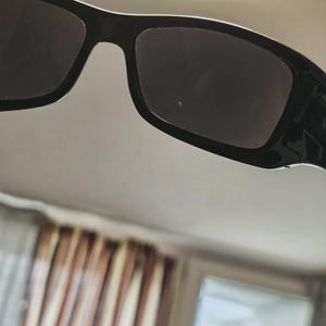K&ouml;nnte beinhalten: Nahaufnahme einer schwarzen Sonnenbrille mit dunklen Gl&auml;sern. Die Sonnenbrille hat einen rechteckigen Rahmen und eine gl&auml;nzende Oberfl&auml;che. Das Bild ist aus einem leicht nach oben gerichteten Winkel aufgenommen, der die Oberseite des Rahmens und der Gl&auml;ser zeigt.