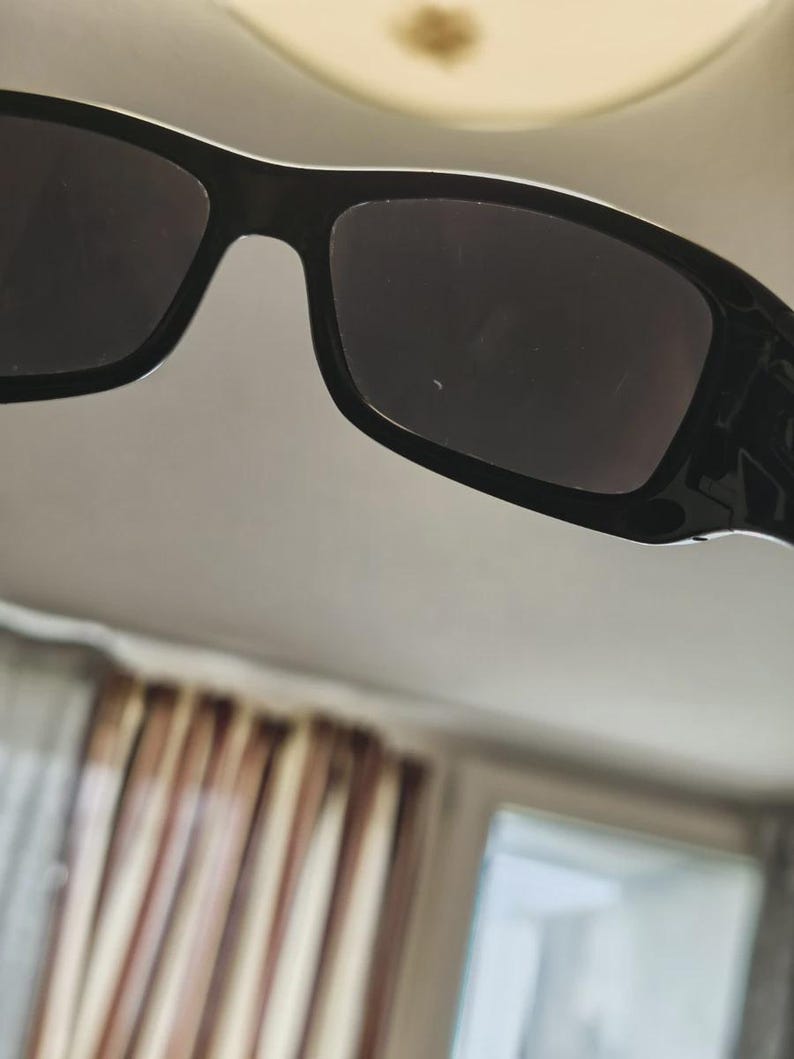 K&ouml;nnte beinhalten: Nahaufnahme einer schwarzen Sonnenbrille mit dunklen Gl&auml;sern. Die Sonnenbrille hat einen rechteckigen Rahmen und eine gl&auml;nzende Oberfl&auml;che. Das Bild ist aus einem leicht nach oben gerichteten Winkel aufgenommen, der die Oberseite des Rahmens und der Gl&auml;ser zeigt.