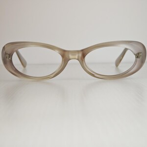 May include: A pair of oval-shaped eyeglasses with a translucent, light taupe-colored frame. The frames feature a smooth, rounded design with small metallic accents at the outer corners where the temples attach to the front of the glasses.