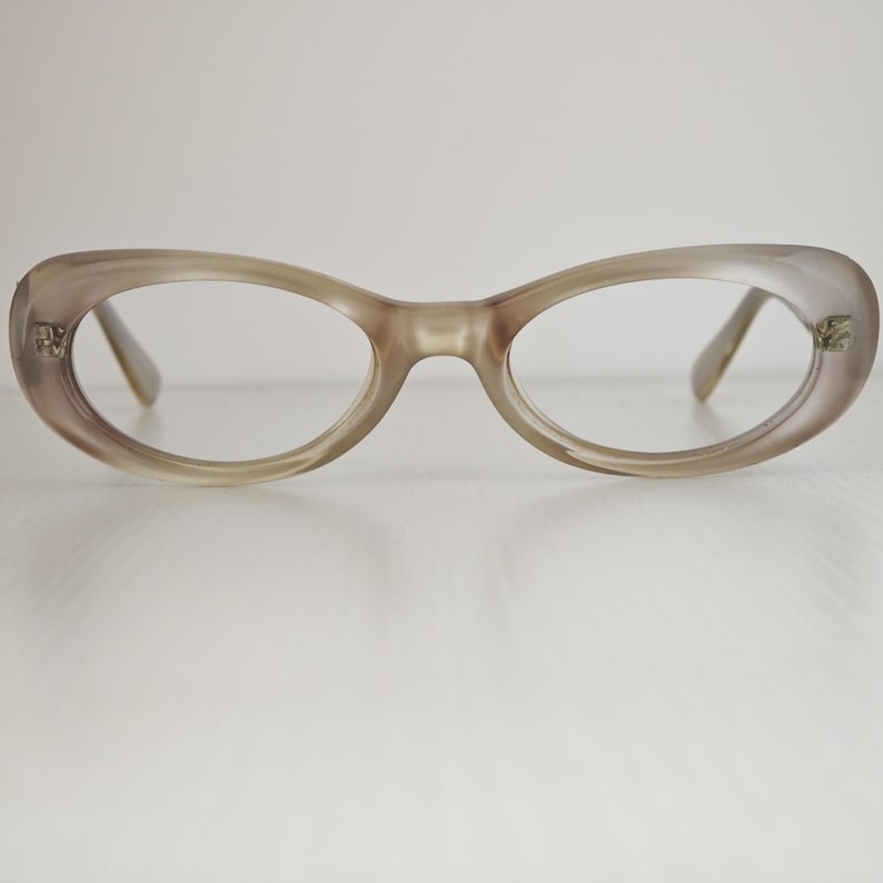 May include: A pair of oval-shaped eyeglasses with a translucent, light taupe-colored frame. The frames feature a smooth, rounded design with small metallic accents at the outer corners where the temples attach to the front of the glasses.