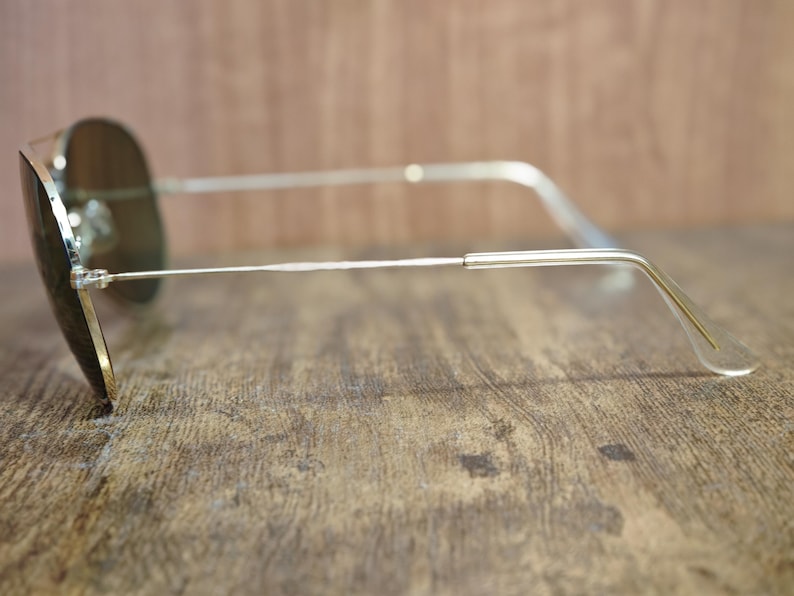 May include: Aviator-style sunglasses with a silver metal frame and dark lenses. The sunglasses are resting on a wooden surface, showcasing the sleek design and classic shape. The lenses appear to be tinted, offering a stylish look.