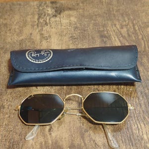 May include: Gold-framed sunglasses with black lenses and a matching black case. The sunglasses have an octagonal shape and clear arms. The case is dark blue with the Ray-Ban logo.