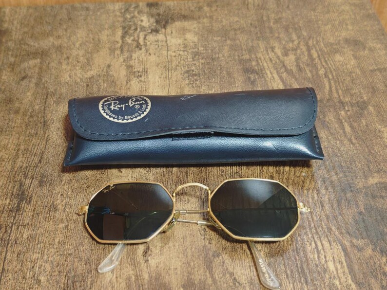 May include: Gold-framed sunglasses with black lenses and a matching black case. The sunglasses have an octagonal shape and clear arms. The case is dark blue with the Ray-Ban logo.
