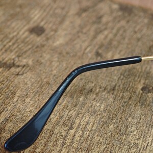 Peut inclure: Gros plan d'une branche de lunettes noire avec une finition brillante. Le bras est l&eacute;g&egrave;rement incurv&eacute; et se termine par une extr&eacute;mit&eacute; arrondie. L'arri&egrave;re-plan est une surface en bois textur&eacute;e.