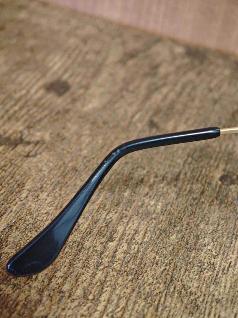 Peut inclure: Gros plan d'une branche de lunettes noire avec une finition brillante. Le bras est l&eacute;g&egrave;rement incurv&eacute; et se termine par une extr&eacute;mit&eacute; arrondie. L'arri&egrave;re-plan est une surface en bois textur&eacute;e.