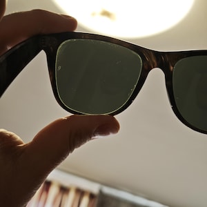 Puede incluir: Gafas de sol con lentes verde oscuro y montura carey marr&oacute;n. Las gafas de sol se sostienen contra una luz brillante, mostrando la forma y el estilo de las gafas. La montura tiene una forma rectangular cl&aacute;sica.