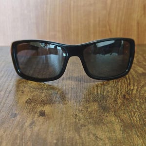K&ouml;nnte beinhalten: Schwarze Sonnenbrille mit dunklen Gl&auml;sern. Die Sonnenbrille hat eine rechteckige Form mit abgerundeten Kanten und einem dicken Rahmen. Die Gl&auml;ser reflektieren die Umgebung. Die Sonnenbrille liegt auf einer Holzoberfl&auml;che.