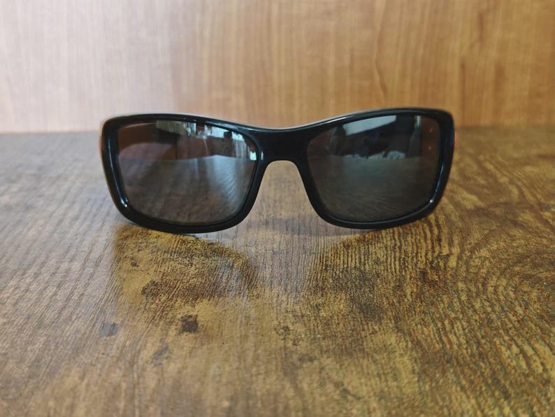 K&ouml;nnte beinhalten: Schwarze Sonnenbrille mit dunklen Gl&auml;sern. Die Sonnenbrille hat eine rechteckige Form mit abgerundeten Kanten und einem dicken Rahmen. Die Gl&auml;ser reflektieren die Umgebung. Die Sonnenbrille liegt auf einer Holzoberfl&auml;che.