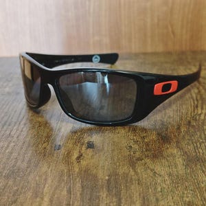 K&ouml;nnte beinhalten: Schwarze Sonnenbrille mit rechteckigem Rahmen und dunklen Gl&auml;sern. Die Sonnenbrille hat ein rotes Logo am B&uuml;gel. Die Sonnenbrille liegt auf einer Holzoberfl&auml;che.