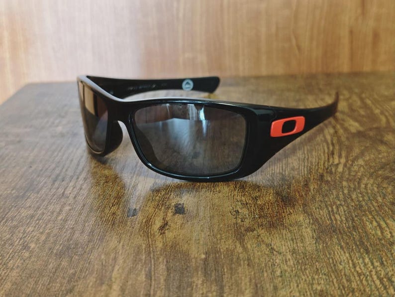 K&ouml;nnte beinhalten: Schwarze Sonnenbrille mit rechteckigem Rahmen und dunklen Gl&auml;sern. Die Sonnenbrille hat ein rotes Logo am B&uuml;gel. Die Sonnenbrille liegt auf einer Holzoberfl&auml;che.