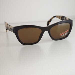 May include: Sunglasses with dark brown frames and brown lenses. The temples feature a cream and black tortoiseshell pattern. A small sticker on the right lens displays the Prada logo with the text 100% U.V. PROTECTION POLARIZED.