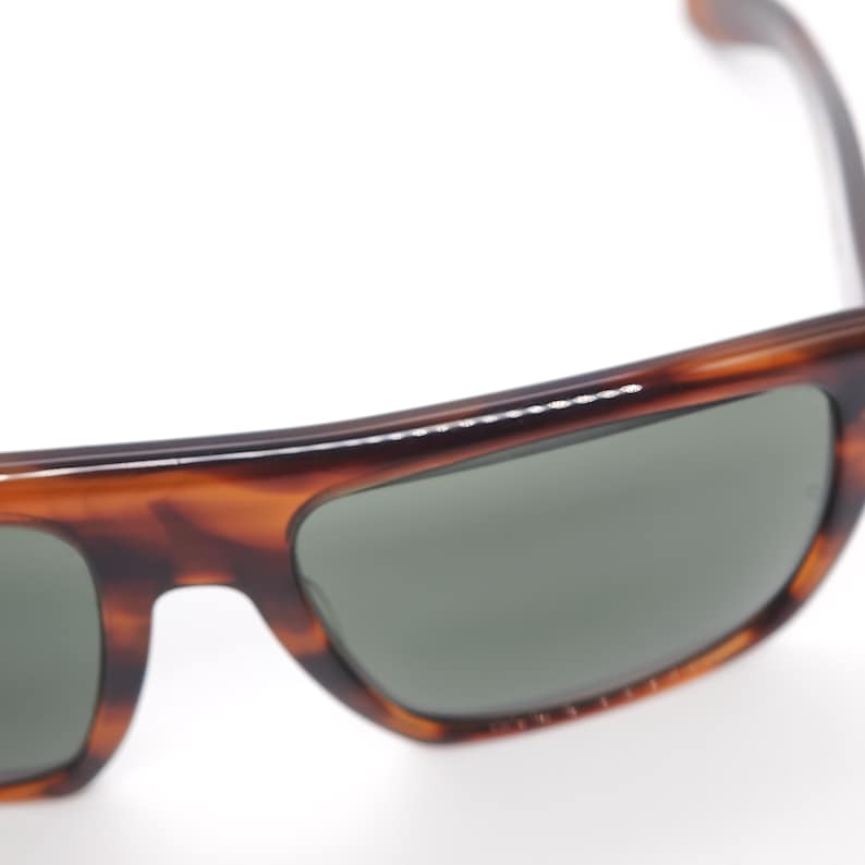 May include: Close-up of sunglasses with a tortoiseshell frame and dark green lenses. The sunglasses have a classic, rectangular shape and a glossy finish. The frame features a brown and amber pattern.