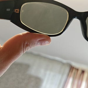 Może przedstawiać: Czarne okulary przeciwsłoneczne z prostokątną oprawą i zielonymi soczewkami. Na lewym zausznika okular&oacute;w przeciwsłonecznych znajduje się napis "W2526". Okulary przeciwsłoneczne są trzymane przez osobę.
