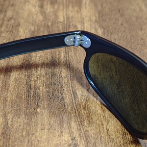 May include: Close-up of black sunglasses with dark lenses. The hinge and arm of the sunglasses are visible, showing the connection point. The lenses are a dark color, and the frame is black.