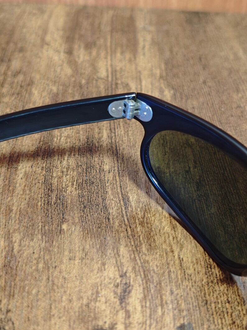 May include: Close-up of black sunglasses with dark lenses. The hinge and arm of the sunglasses are visible, showing the connection point. The lenses are a dark color, and the frame is black.