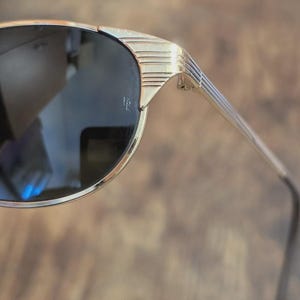 May include: Gold-toned sunglasses with dark lenses. The frames have a sleek, geometric design with horizontal lines on the temples. The sunglasses have a classic, retro aesthetic.