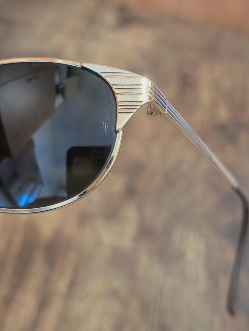 May include: Gold-toned sunglasses with dark lenses. The frames have a sleek, geometric design with horizontal lines on the temples. The sunglasses have a classic, retro aesthetic.
