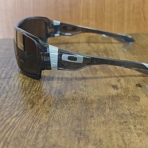 May include: Black rectangular sunglasses with a silver accent on the temples and arms. The lenses are dark, and the frame is a translucent black. The Oakley logo is visible on the arm.
