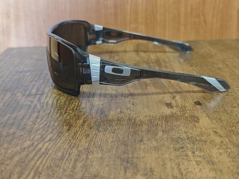 May include: Black rectangular sunglasses with a silver accent on the temples and arms. The lenses are dark, and the frame is a translucent black. The Oakley logo is visible on the arm.
