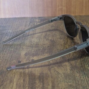 May include: A pair of gray sunglasses with dark lenses, resting on a wooden surface. The sunglasses have a sleek, modern design with thin temples and a subtle logo on the side. The lenses appear to be tinted, offering protection from the sun.
