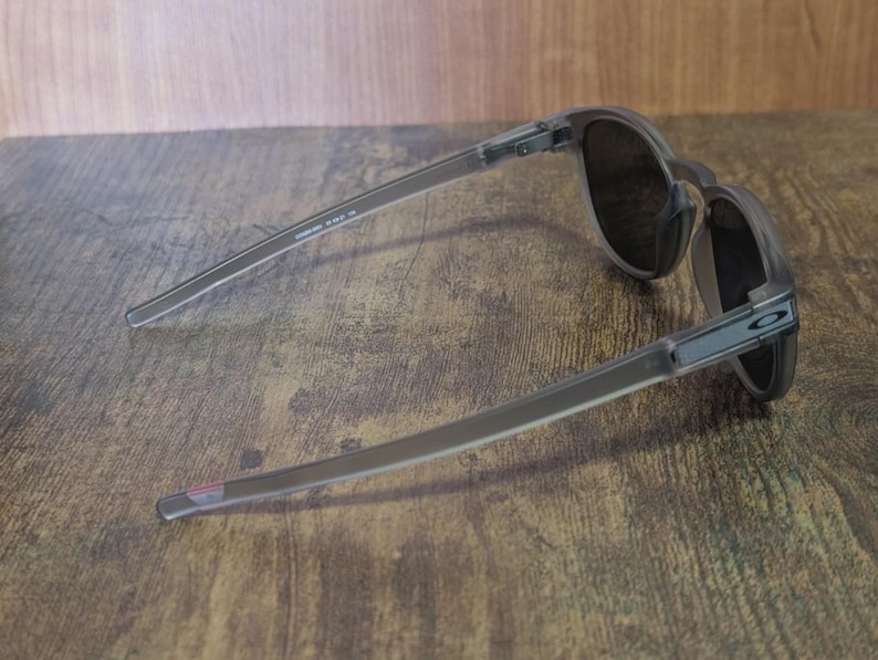 May include: A pair of gray sunglasses with dark lenses, resting on a wooden surface. The sunglasses have a sleek, modern design with thin temples and a subtle logo on the side. The lenses appear to be tinted, offering protection from the sun.