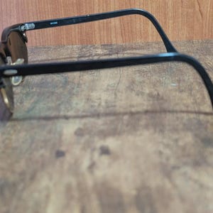 May include: Black-framed sunglasses with dark lenses. The glasses have a classic design with a browline and a curved temple arm. The sunglasses are set against a wooden background.