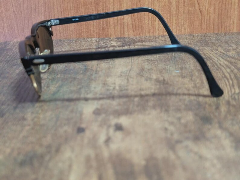 May include: Black-framed sunglasses with dark lenses. The glasses have a classic design with a browline and a curved temple arm. The sunglasses are set against a wooden background.