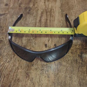 May include: Black sunglasses with a dark lens, measured at approximately 5.5 inches across the frame. The sunglasses are placed on a wooden surface, with a yellow measuring tape extending across the frame.