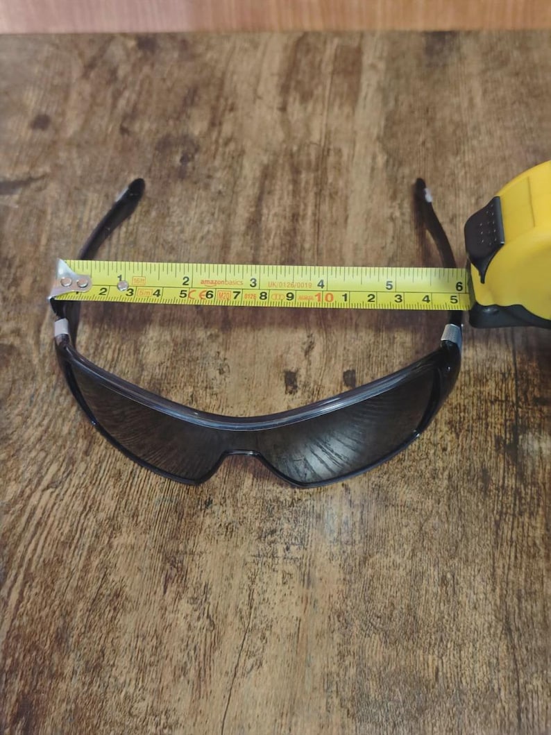 May include: Black sunglasses with a dark lens, measured at approximately 5.5 inches across the frame. The sunglasses are placed on a wooden surface, with a yellow measuring tape extending across the frame.