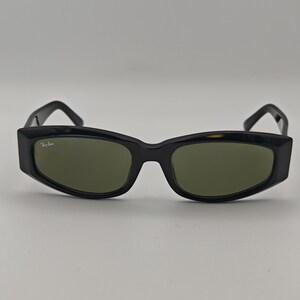 Może przedstawiać: Czarne okulary przeciwsłoneczne z prostokątną oprawką i zielonymi soczewkami. Okulary mają grubą oprawkę, a logo Ray-Ban jest widoczne po lewej stronie.