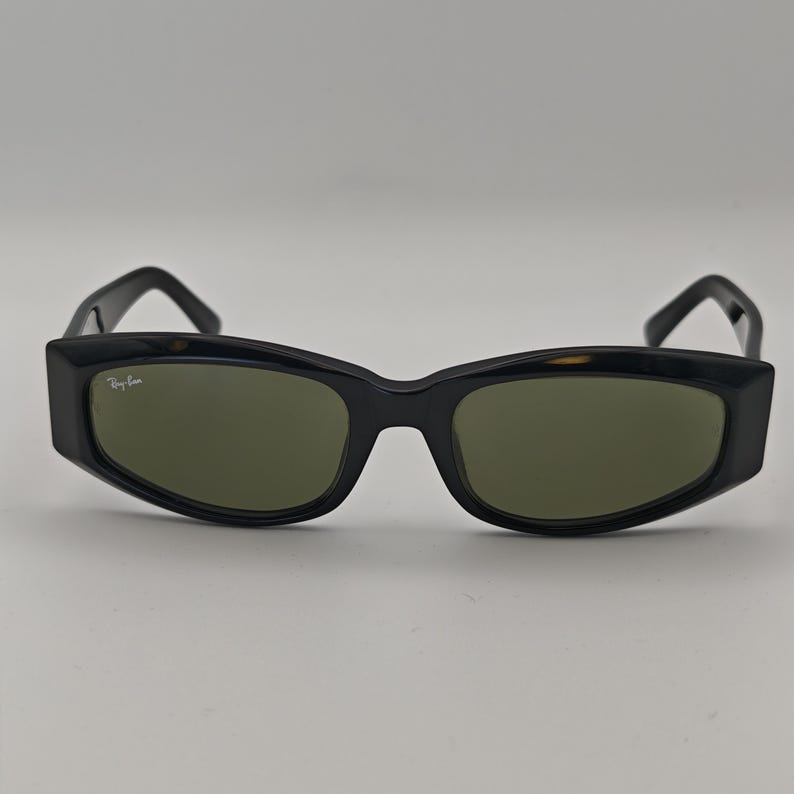 Może przedstawiać: Czarne okulary przeciwsłoneczne z prostokątną oprawką i zielonymi soczewkami. Okulary mają grubą oprawkę, a logo Ray-Ban jest widoczne po lewej stronie.