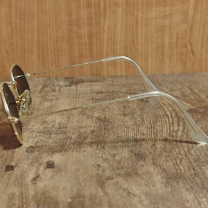 May include: A pair of sunglasses with a gold-colored frame and brown lenses. The glasses are resting on a wooden surface. The temples are silver and curve at the ear pieces.