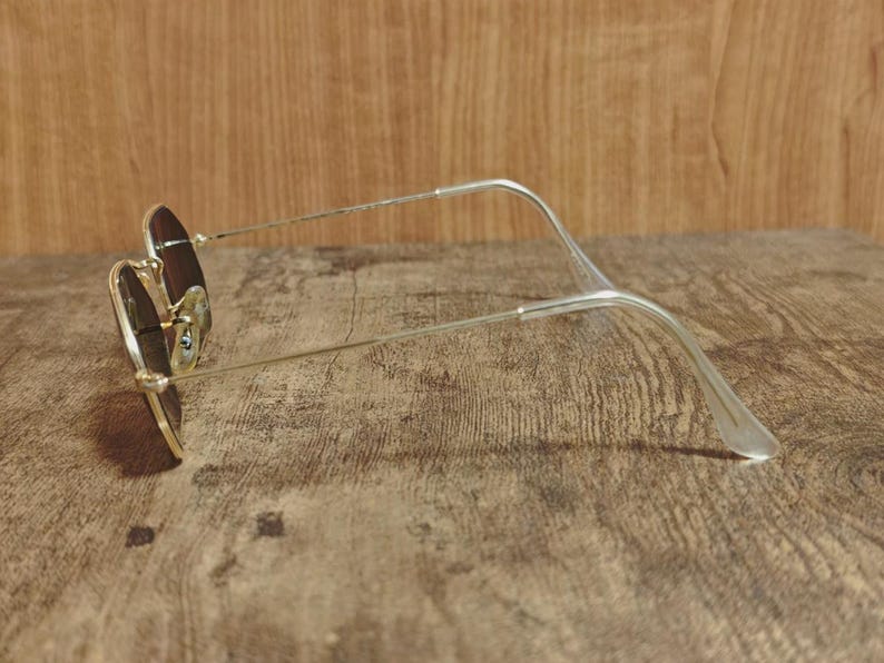 May include: A pair of sunglasses with a gold-colored frame and brown lenses. The glasses are resting on a wooden surface. The temples are silver and curve at the ear pieces.