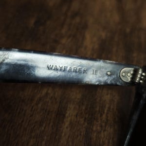 Puede incluir: Primer plano de una patilla de gafas de sol Wayfarer II plateada. El texto "WAYFARER II" est&aacute; grabado en la patilla met&aacute;lica. La patilla est&aacute; unida a la bisagra y la montura es visible.