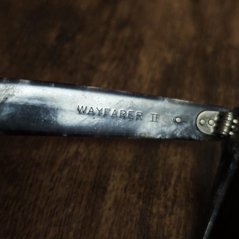 Puede incluir: Primer plano de una patilla de gafas de sol Wayfarer II plateada. El texto "WAYFARER II" est&aacute; grabado en la patilla met&aacute;lica. La patilla est&aacute; unida a la bisagra y la montura es visible.
