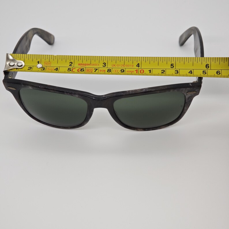 Puede incluir: Gafas de sol negras con lentes verde oscuro. La montura tiene un patr&oacute;n moteado. Una cinta m&eacute;trica amarilla muestra que las gafas de sol miden aproximadamente 14,6 cm de ancho.