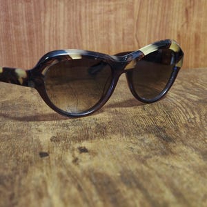 May include: A pair of cat-eye sunglasses with a tortoiseshell pattern on the frames and arms. The lenses are a gradient brown color. The sunglasses are resting on a wooden surface.