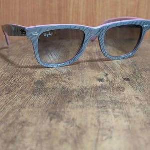 Puede incluir: Gafas de sol grises y rosas con un patr&oacute;n de vetas de madera. Las gafas de sol tienen lentes marrones y el logotipo de Ray-Ban es visible en la lente derecha. Las patillas de las gafas son rosas.