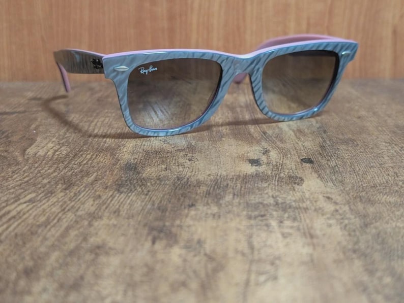Puede incluir: Gafas de sol grises y rosas con un patr&oacute;n de vetas de madera. Las gafas de sol tienen lentes marrones y el logotipo de Ray-Ban es visible en la lente derecha. Las patillas de las gafas son rosas.