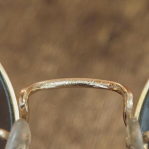 May include: Close-up of gold-toned Ray-Ban sunglasses. The bridge of the glasses features the brand name and "USA" etched into the metal. The lenses are dark, and the nose pads are clear.