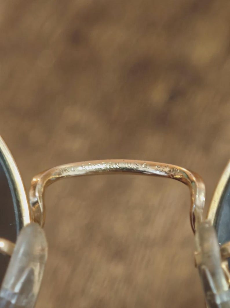 May include: Close-up of gold-toned Ray-Ban sunglasses. The bridge of the glasses features the brand name and "USA" etched into the metal. The lenses are dark, and the nose pads are clear.