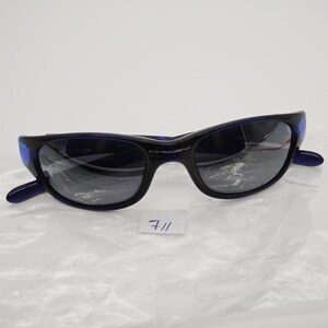May include: Black and blue sunglasses with a curved frame and dark lenses. The sunglasses have a sporty design and the brand name "Oakley" is visible on the frame. A small white tag with the number "711" is also present.