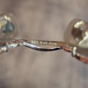 Peut inclure: Gros plan d'une monture de lunettes en m&eacute;tal argent&eacute; avec le texte "B&L RAY-BAN" grav&eacute; sur le pont. La monture a des verres transparents et un design incurv&eacute;. L'arri&egrave;re-plan est flou.