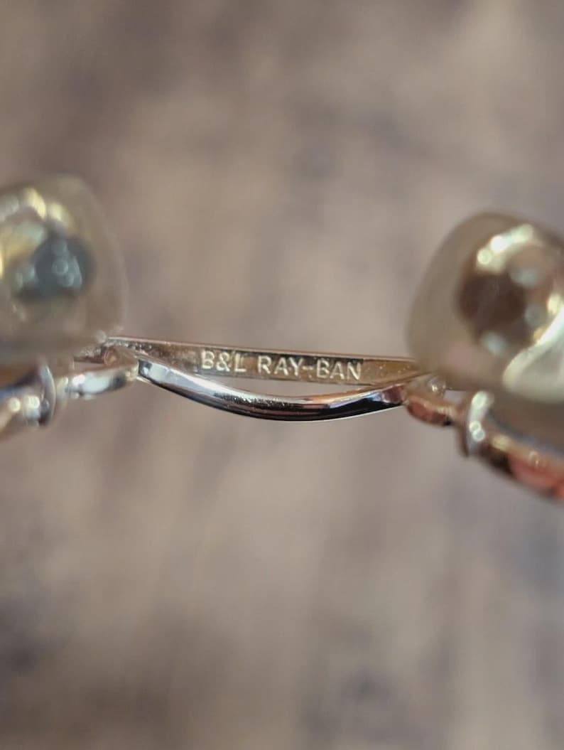 Peut inclure: Gros plan d'une monture de lunettes en m&eacute;tal argent&eacute; avec le texte "B&L RAY-BAN" grav&eacute; sur le pont. La monture a des verres transparents et un design incurv&eacute;. L'arri&egrave;re-plan est flou.