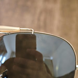 May include: Close-up of aviator sunglasses with gold-colored frames and dark lenses. The lenses reflect the surrounding environment. The top bar and temples are visible, with a light-colored temple tip.